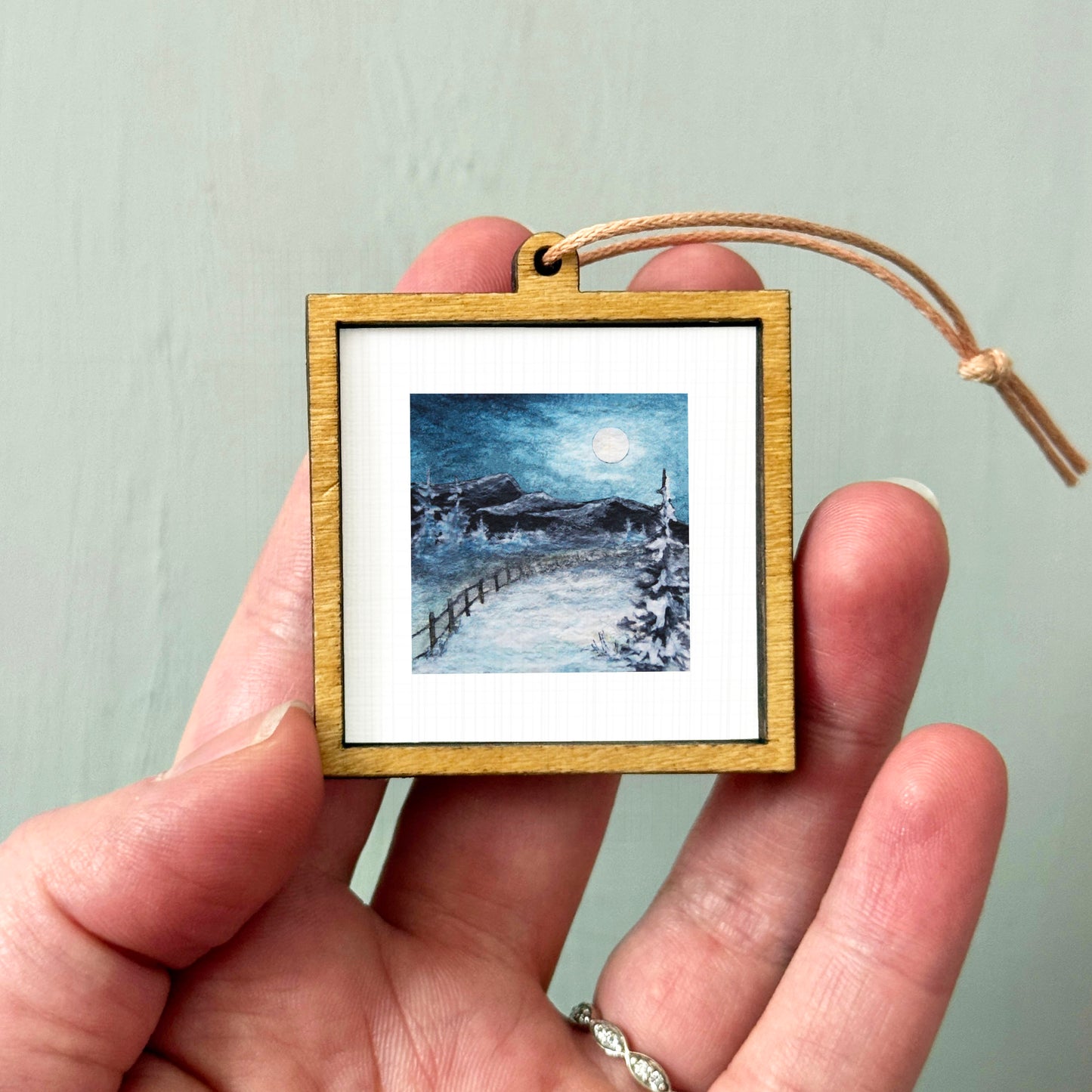 A hand holding a small wooden picture frame containing a painting of a snowy landscape with a full moon in the sky.