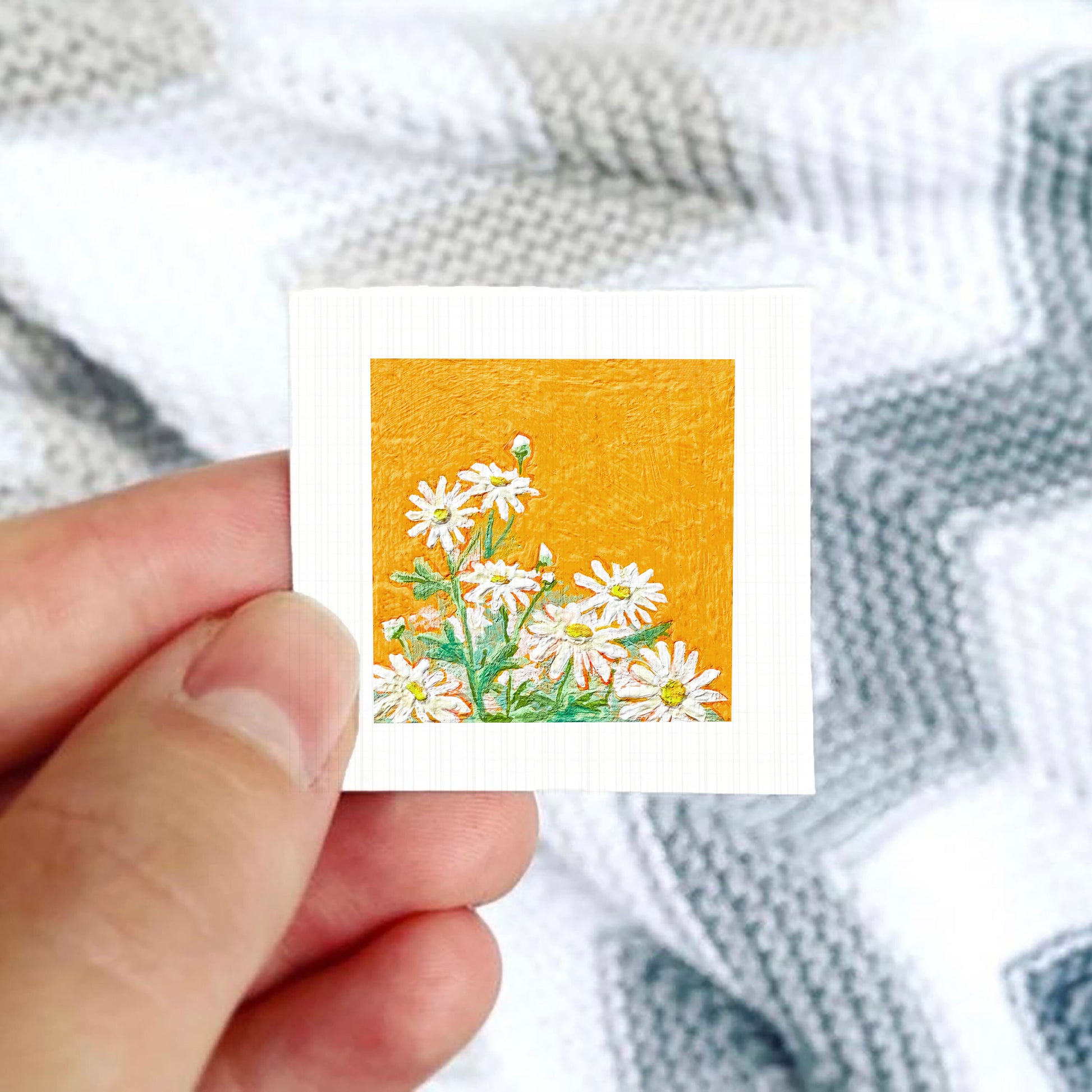 A hand is holding a small square card with a vibrant illustration of white daisies against an orange background.