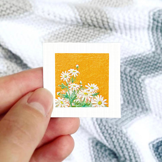 A hand is holding a small square card with a vibrant illustration of white daisies against an orange background.