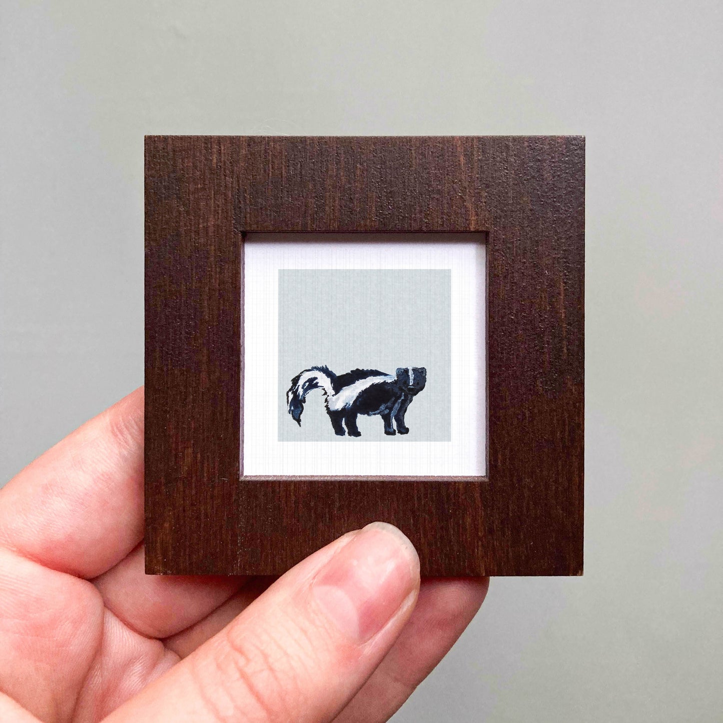 A hand is holding a small wooden frame containing a picture of a skunk.