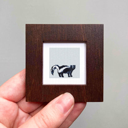 A hand is holding a small wooden frame containing a picture of a skunk.