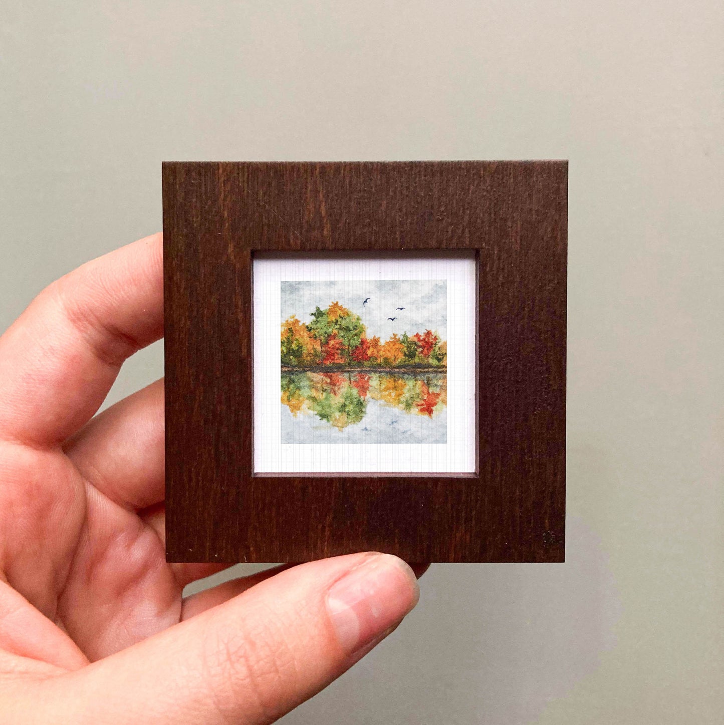 A hand is holding a small, square-shaped wooden frame containing a miniature painting of a landscape with trees and a lake.