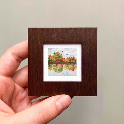 A hand is holding a small, square-shaped wooden frame containing a miniature painting of a landscape with trees and a lake.