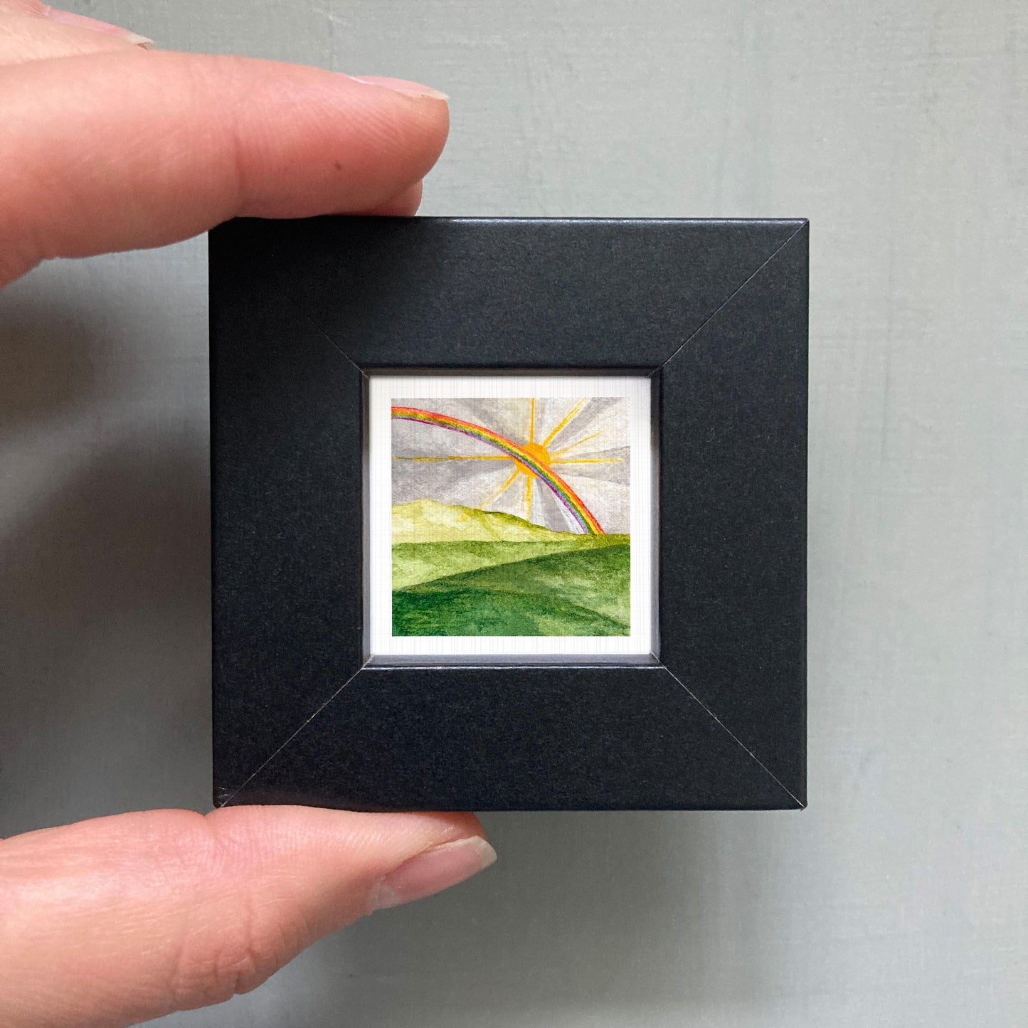 A hand is holding a small black frame containing a watercolor painting of a landscape with a rainbow in the background.