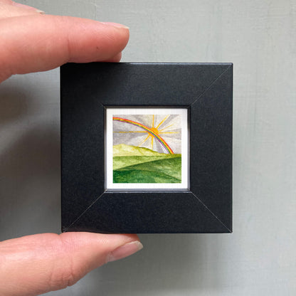 A hand is holding a small black frame containing a watercolor painting of a landscape with a rainbow in the background.
