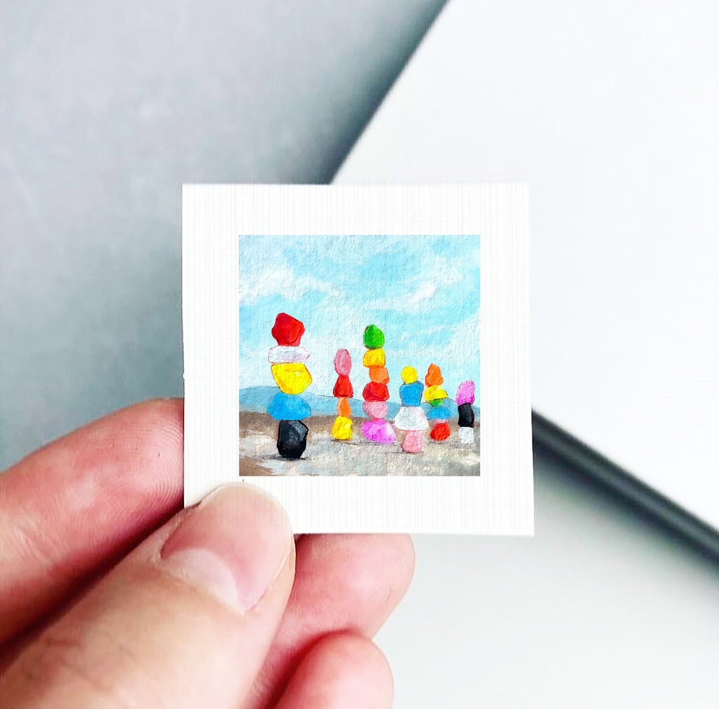 A hand is holding a small, square-shaped painting depicting a beach scene with colorful objects, including a yellow ball, a red ball, and a black ball.
