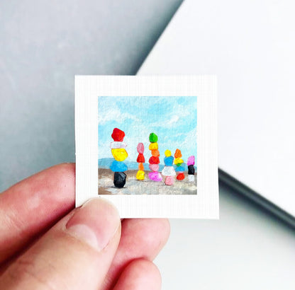 A hand is holding a small, square-shaped painting depicting a beach scene with colorful objects, including a yellow ball, a red ball, and a black ball.