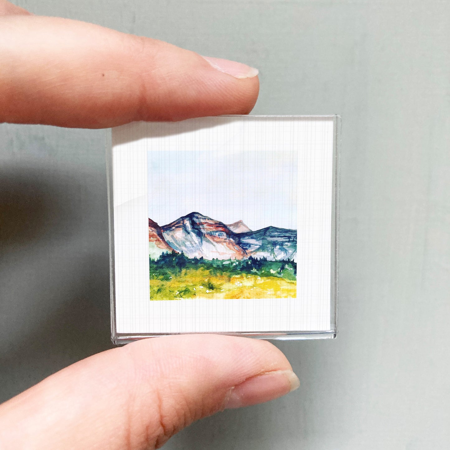 A person's hand holding a small, square-shaped picture of a mountain landscape.