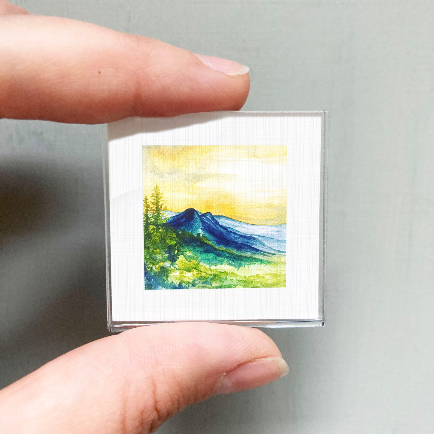 A person's hand holding a small square picture of a mountain landscape with a yellow sky and green trees.