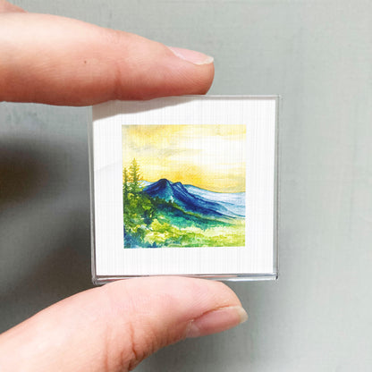 A person's hand holding a small square picture of a mountain landscape with a yellow sky and green trees.