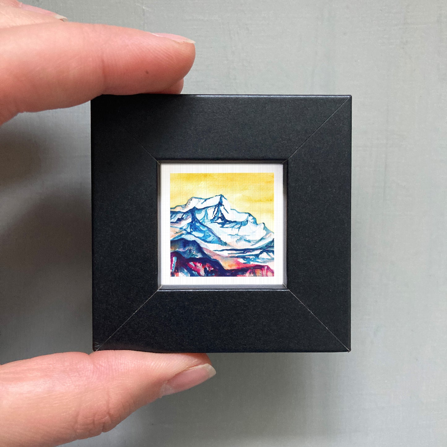A person's hand holding a small, square-shaped black frame containing a watercolor painting of a mountain landscape.