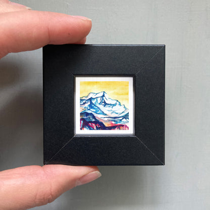A person's hand holding a small, square-shaped black frame containing a watercolor painting of a mountain landscape.