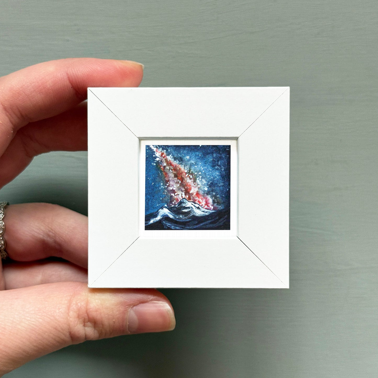 A hand is holding a small white square frame containing a painting of a colorful, abstract underwater scene with a wave-like shape.