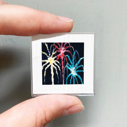 A person's hand is holding a small square picture of colorful fireworks against a gray background.
