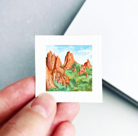 A hand is holding a small square painting depicting a landscape with red mountains and green vegetation.