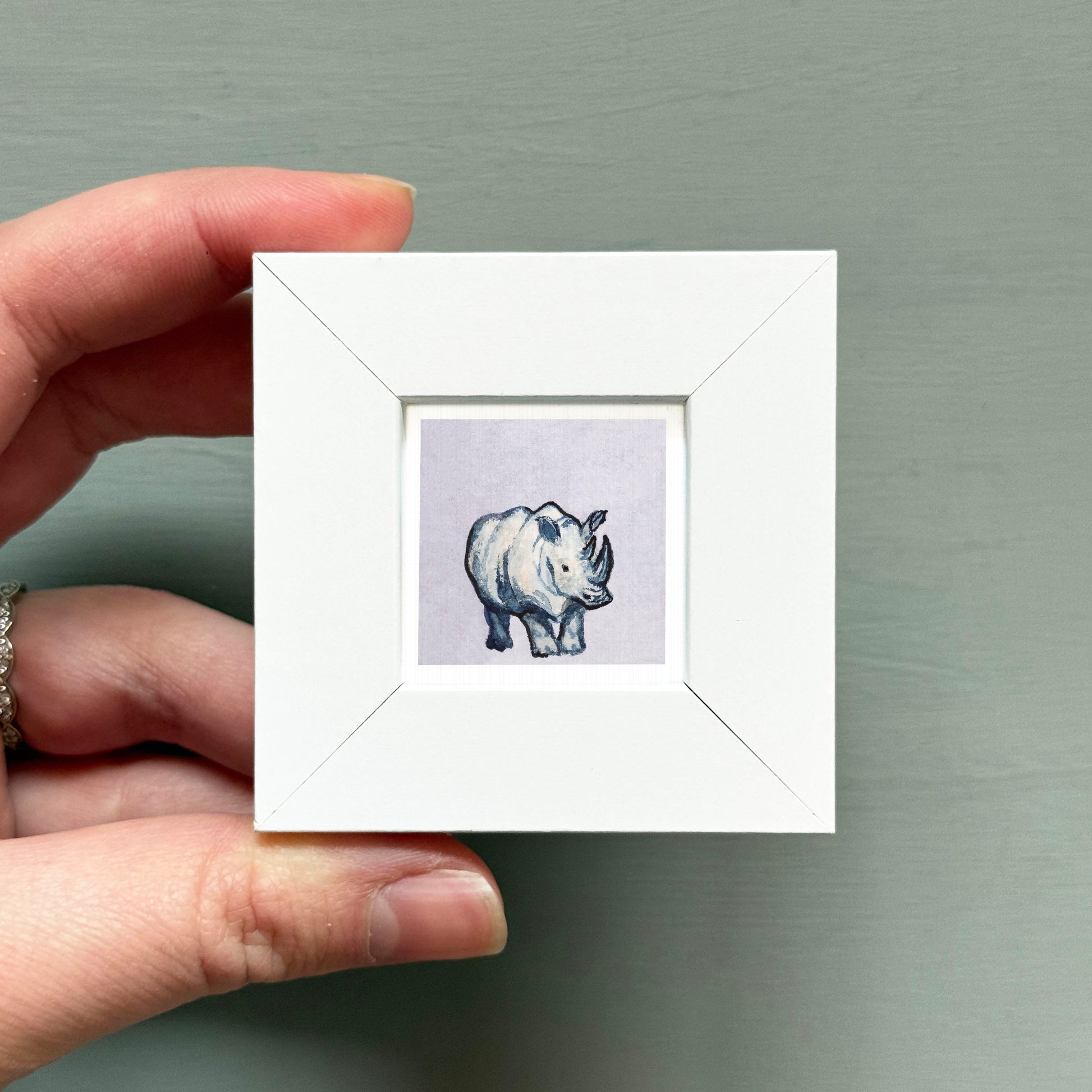 A hand is holding a small white square frame containing a single illustration of a rhinoceros.