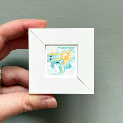 A hand is holding a small white square frame containing a painting of a sun and clouds.