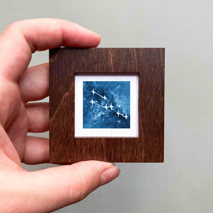 A hand is holding a small wooden frame containing a framed image of a starry night sky.