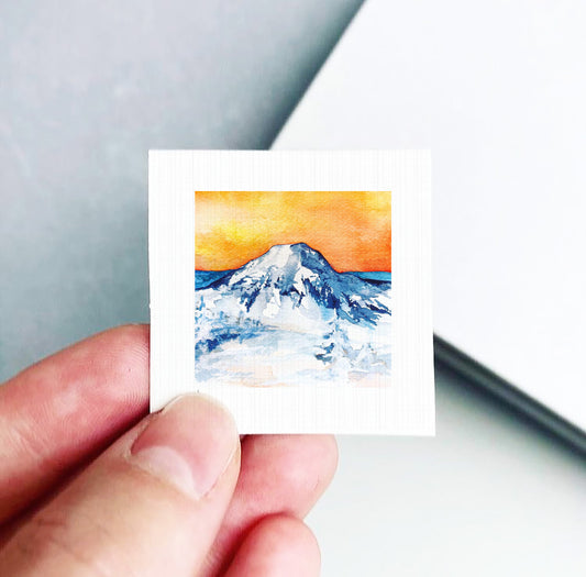 A hand is holding a small square artwork depicting a mountain landscape with a vibrant orange sky.