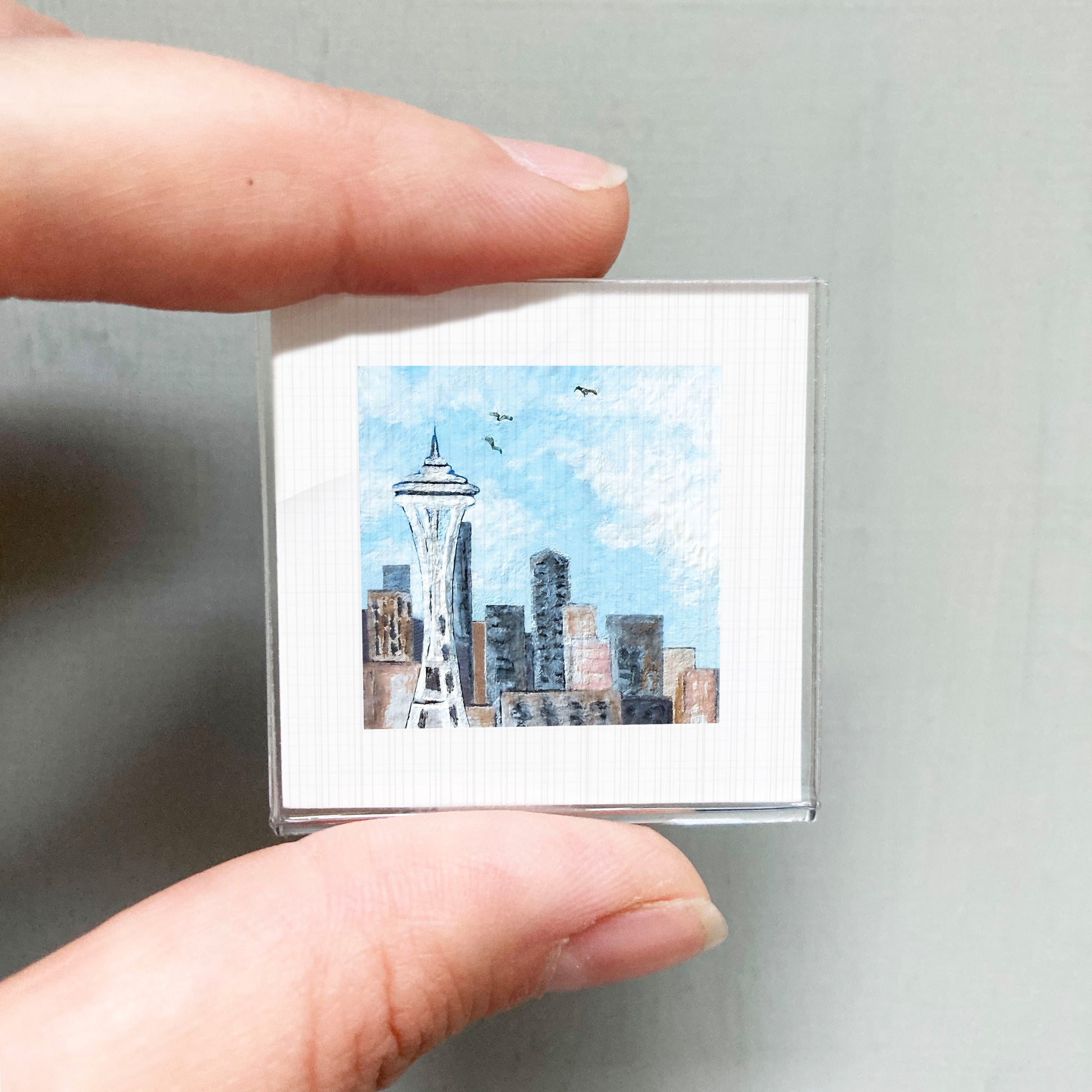A person's hand holding a small square picture of the Seattle Space Needle against a gray background.