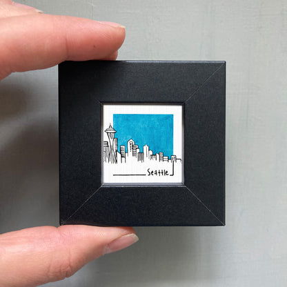 A hand is holding a small square frame containing a drawing of the Seattle skyline.