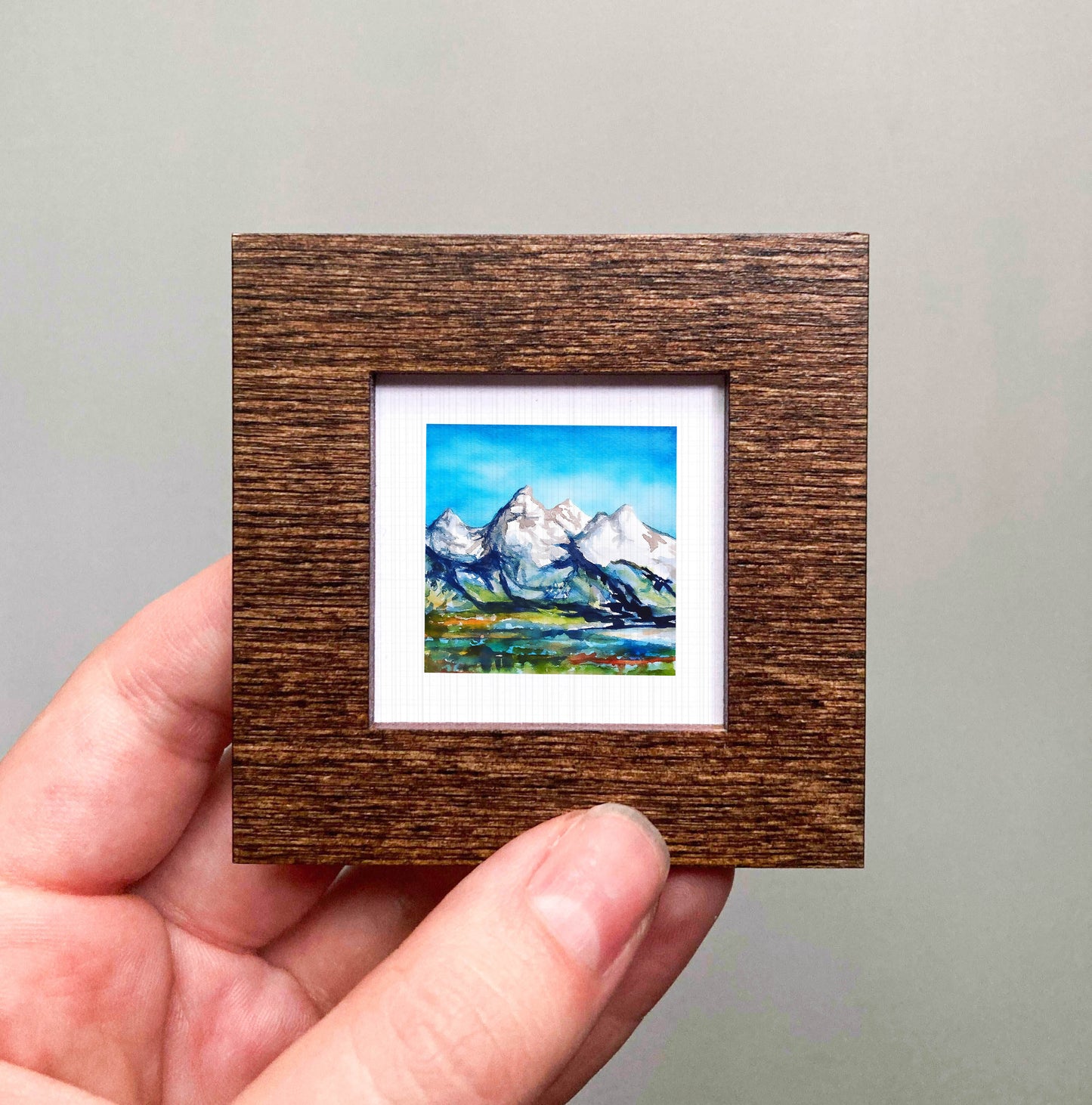 A hand holding a small wooden frame containing a painting of a mountain landscape.