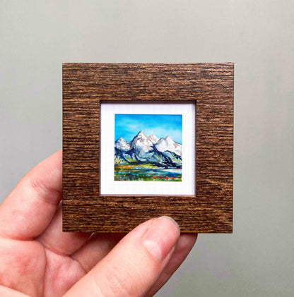 A hand holding a small wooden frame containing a painting of a mountain landscape.