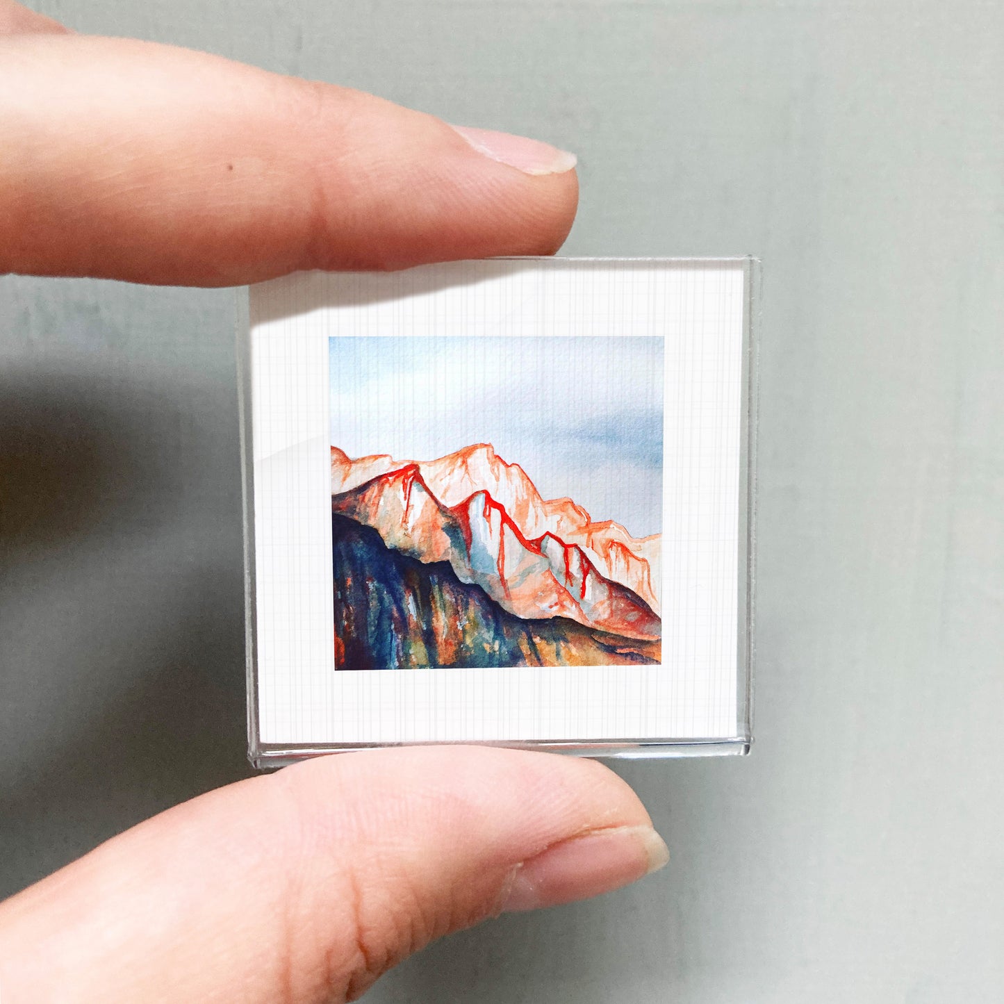 A person's hand holding a small square picture of a mountain landscape with a vibrant, colorful scene.