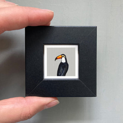 A person's hand is holding a small black square frame containing a framed image of a toucan.