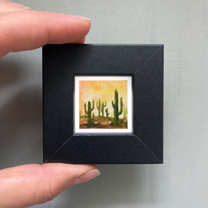 A person's hand holding a small black frame containing a framed painting of cacti.