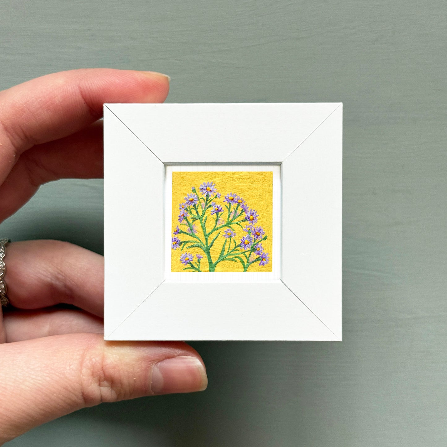 A hand is holding a small white square frame containing a painting of a tree with purple flowers.