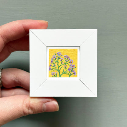 A hand is holding a small white square frame containing a painting of a tree with purple flowers.