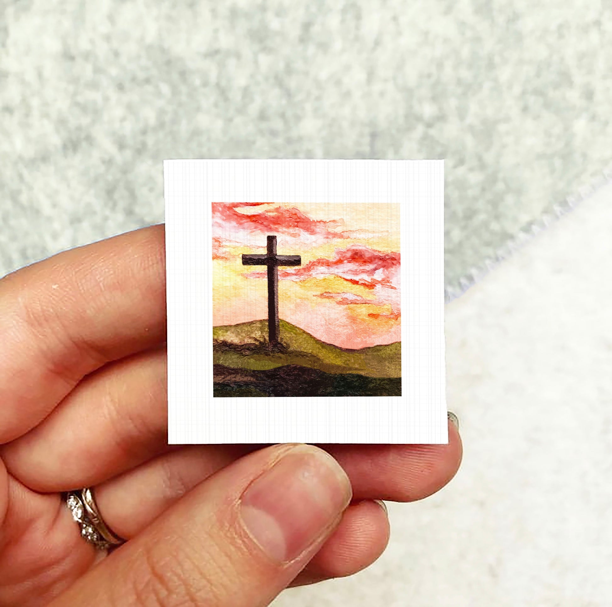 A hand holding a small square card with a painting of a cross against a sunset landscape.