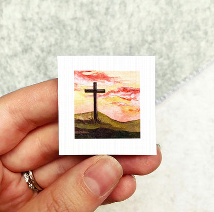 A hand holding a small square card with a painting of a cross against a sunset landscape.