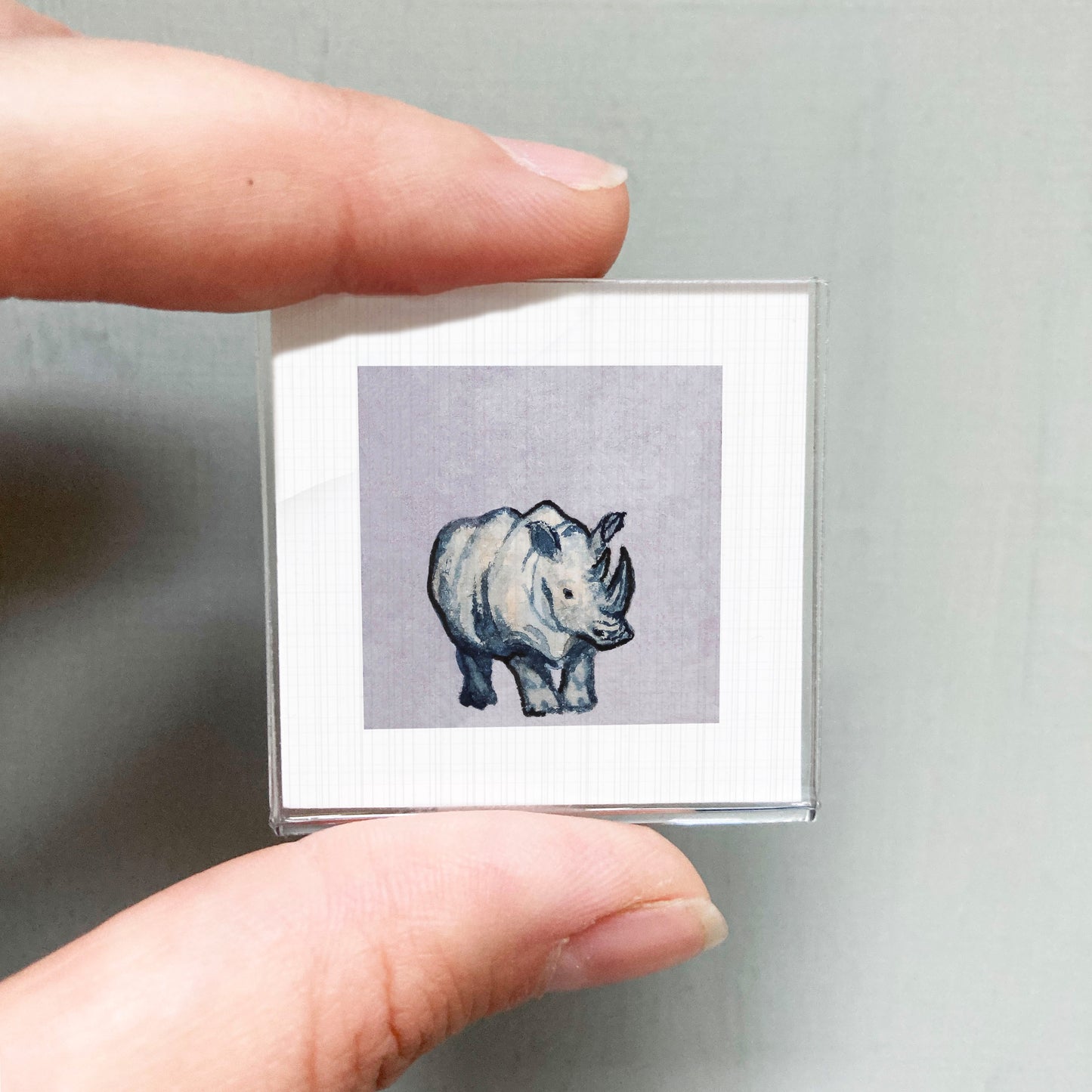 A person's hand is holding a small square picture of a rhinoceros.