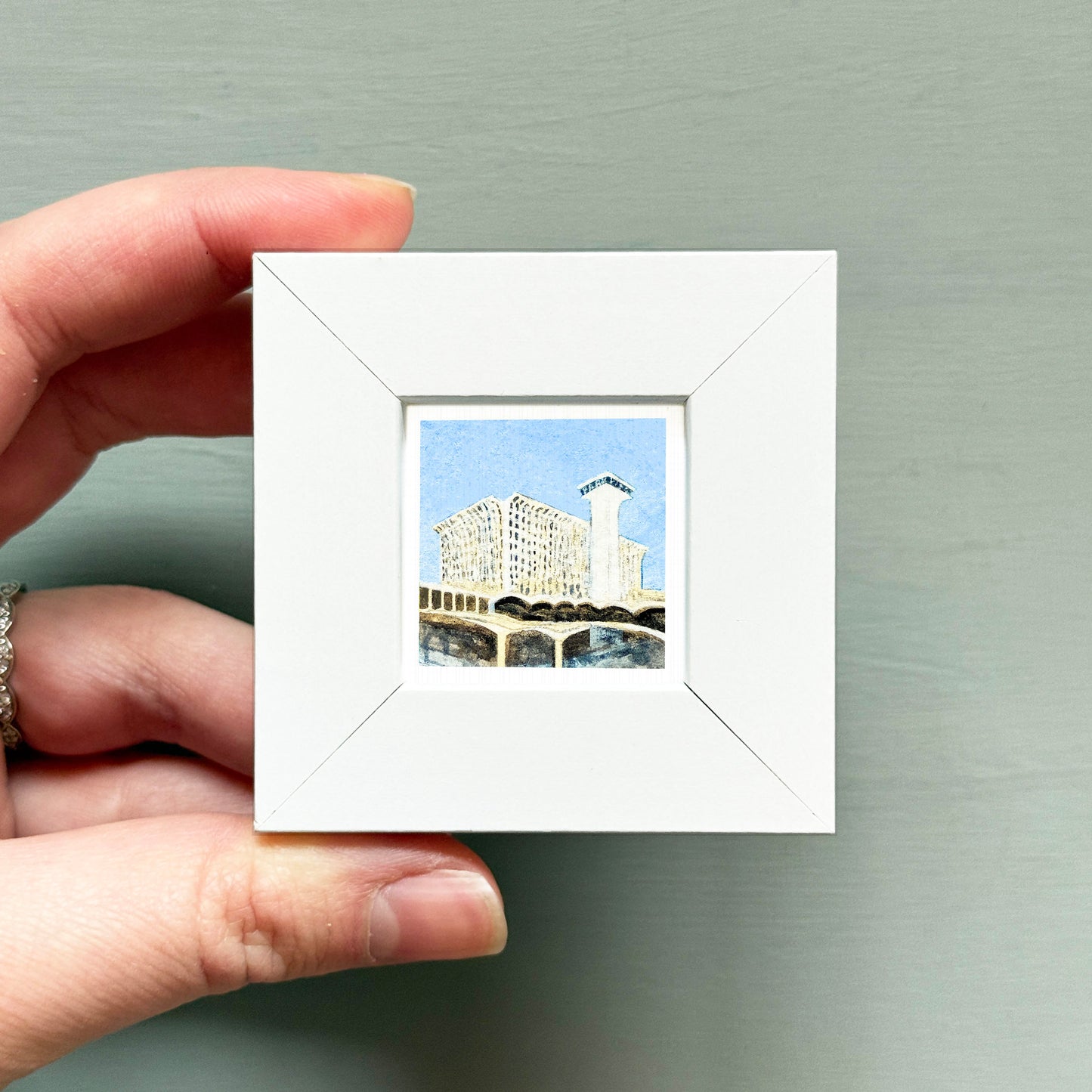 A hand is holding a small white square frame containing a miniature painting of a building.