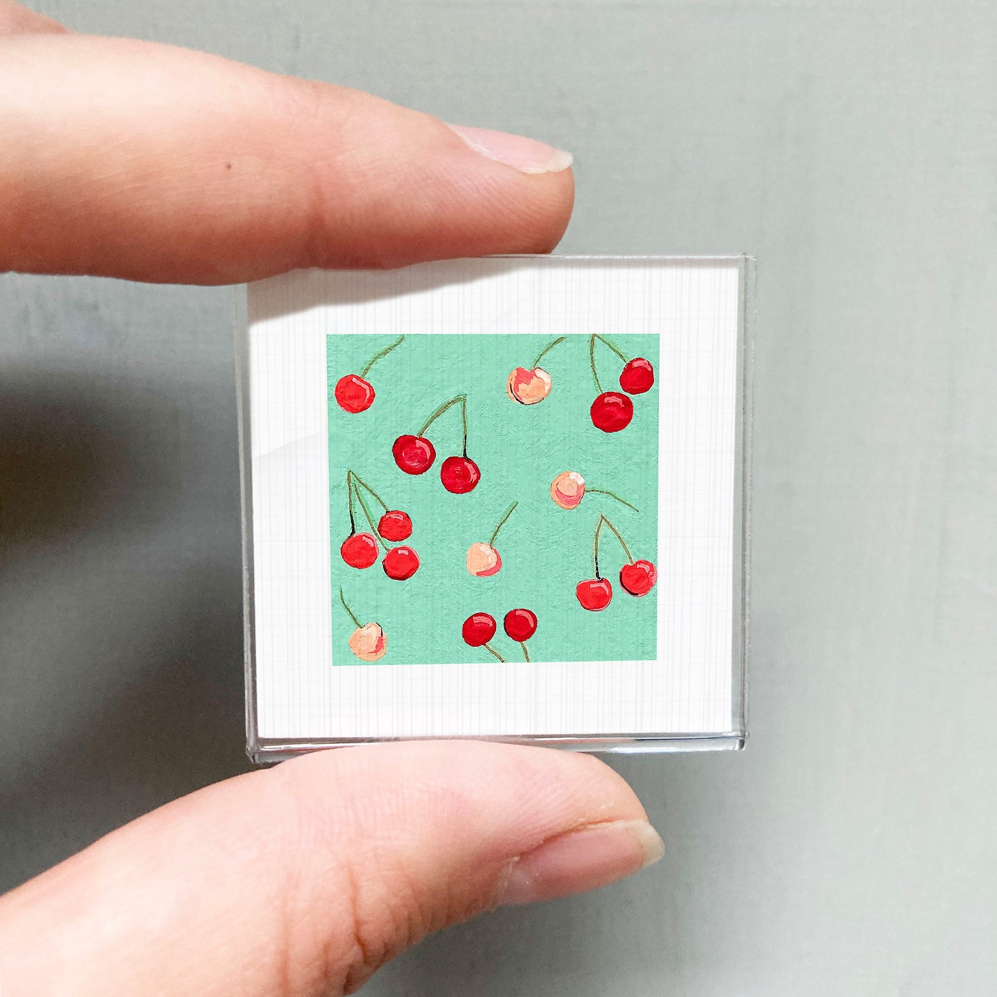 A person's hand is holding a small square with a vibrant illustration of cherries on a green background.