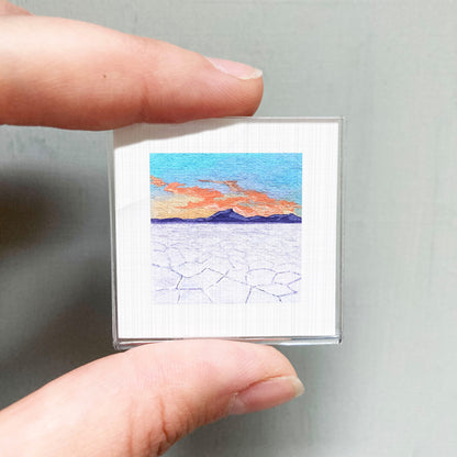 A person's hand is holding a small, square-shaped picture that depicts a landscape with mountains and a colorful sky.