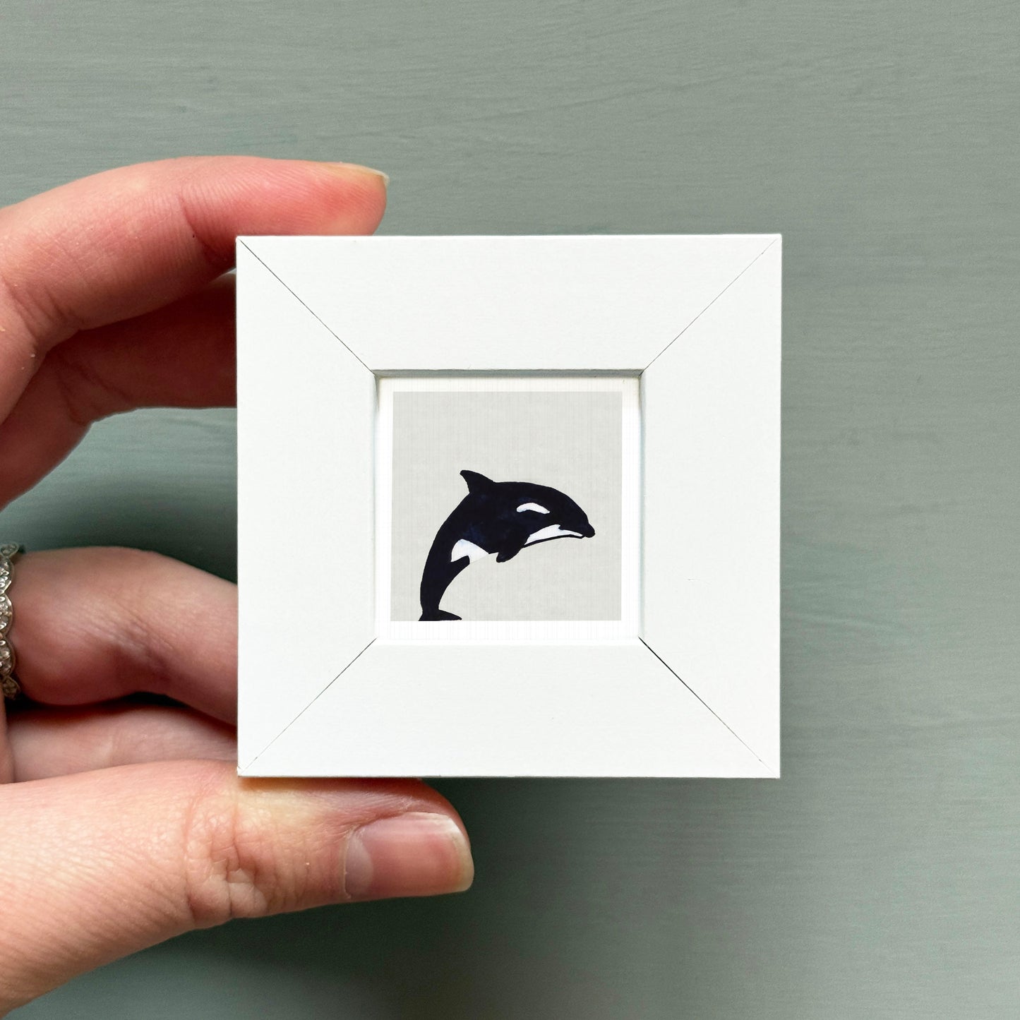 A hand is holding a small white square frame containing a black silhouette of a whale.