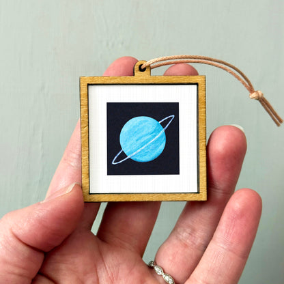 A hand holding a small wooden frame containing a picture of a blue planet with a ring around it.