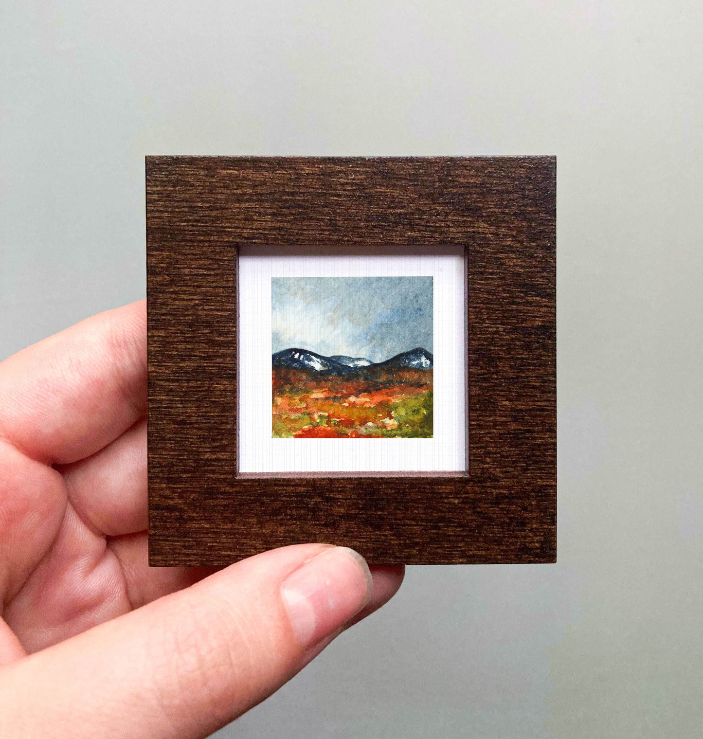 A hand is holding a small, square-shaped wooden frame containing a miniature painting of a landscape with mountains and a field.