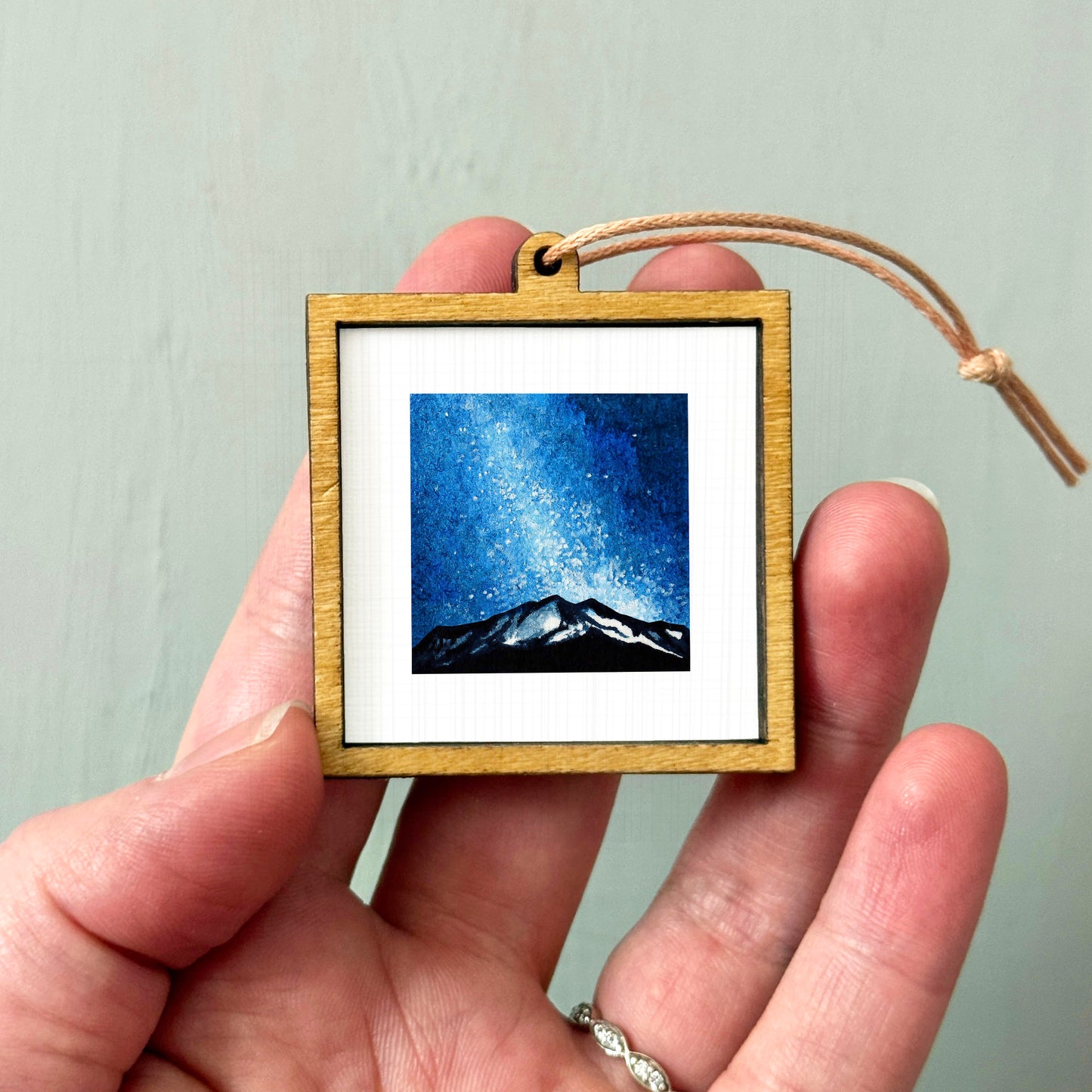 A hand holding a small wooden frame containing a painting of a mountain landscape under a starry night sky.