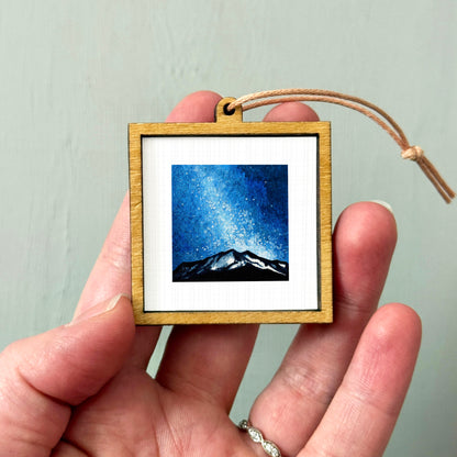 A hand holding a small wooden frame containing a painting of a mountain landscape under a starry night sky.