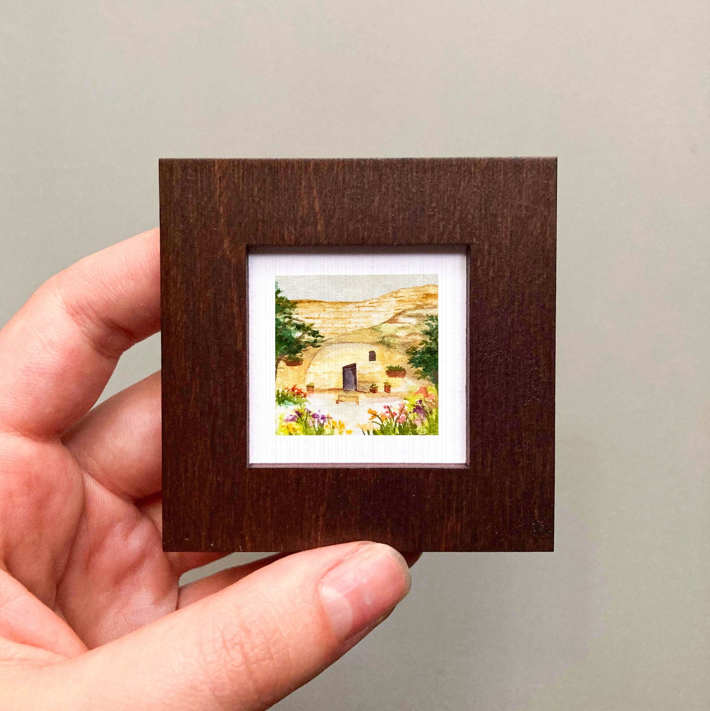 A hand is holding a small, square-shaped wooden frame containing a watercolor painting of a rural landscape with a house and garden.
