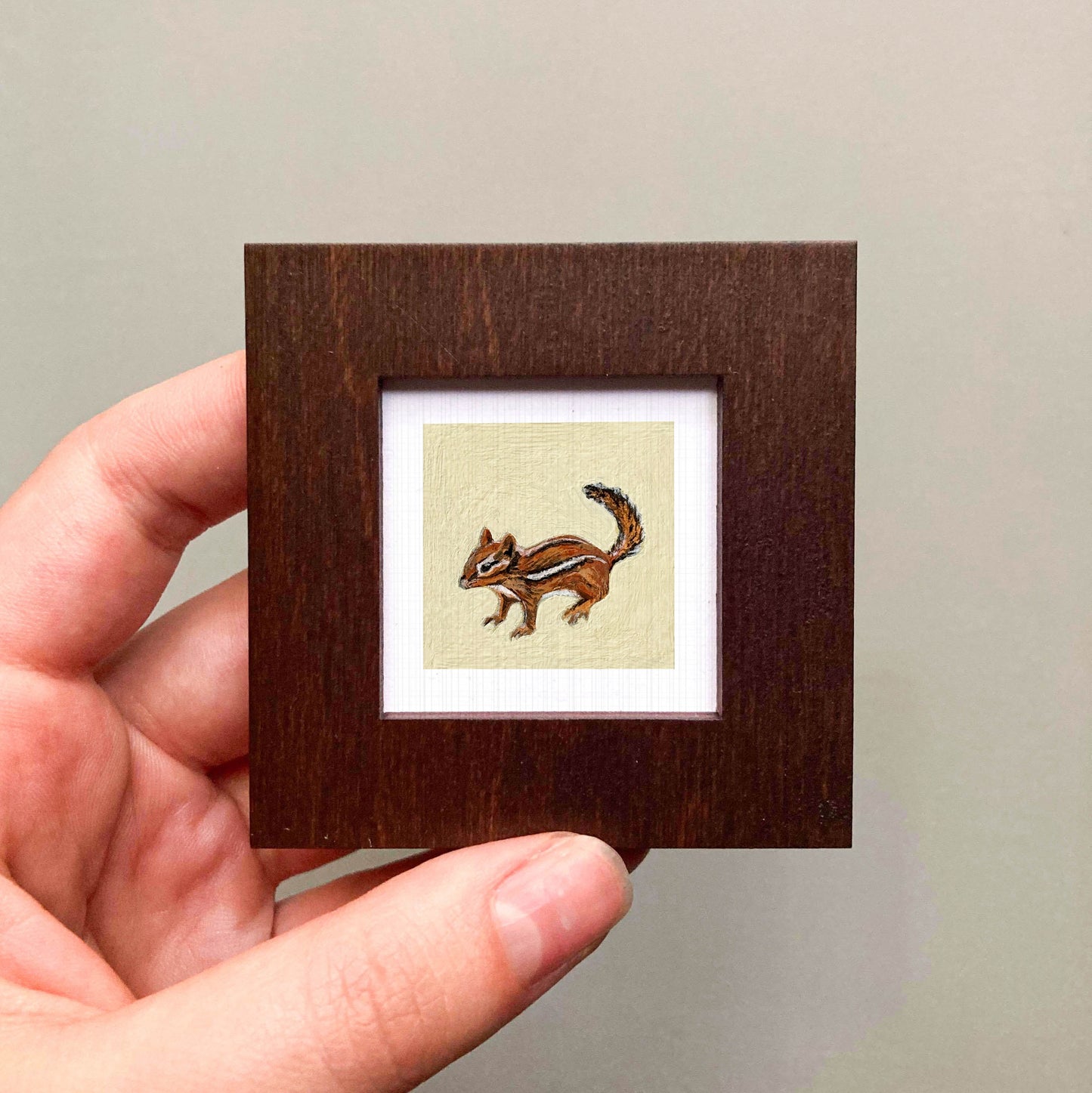 A person's hand is holding a small wooden frame containing a painting of a chipmunk.