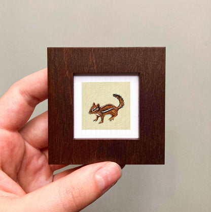 A person's hand is holding a small wooden frame containing a painting of a chipmunk.