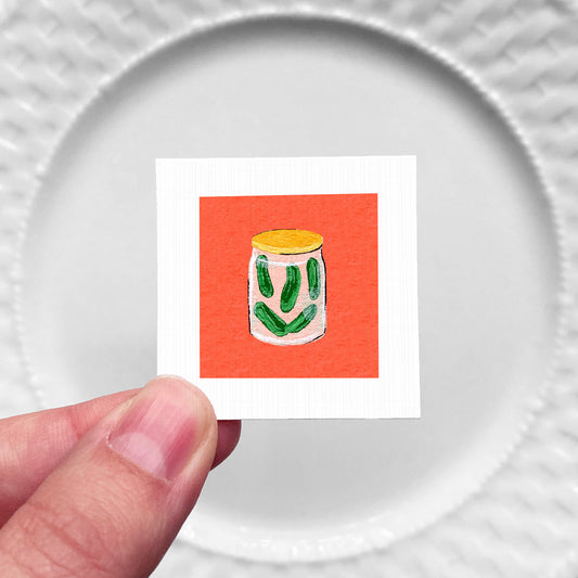 A hand is holding a small square with an illustration of a jar of pickles on it, against a white background.