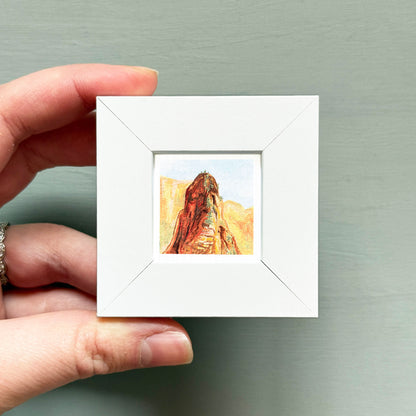 A hand is holding a small white square frame containing a painting of a mountain landscape.