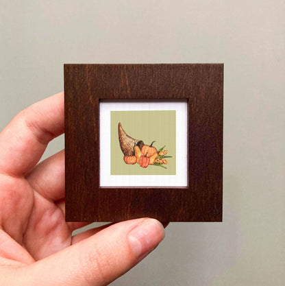 A hand is holding a small wooden frame containing a framed illustration of a cornucopia with various fruits and vegetables.