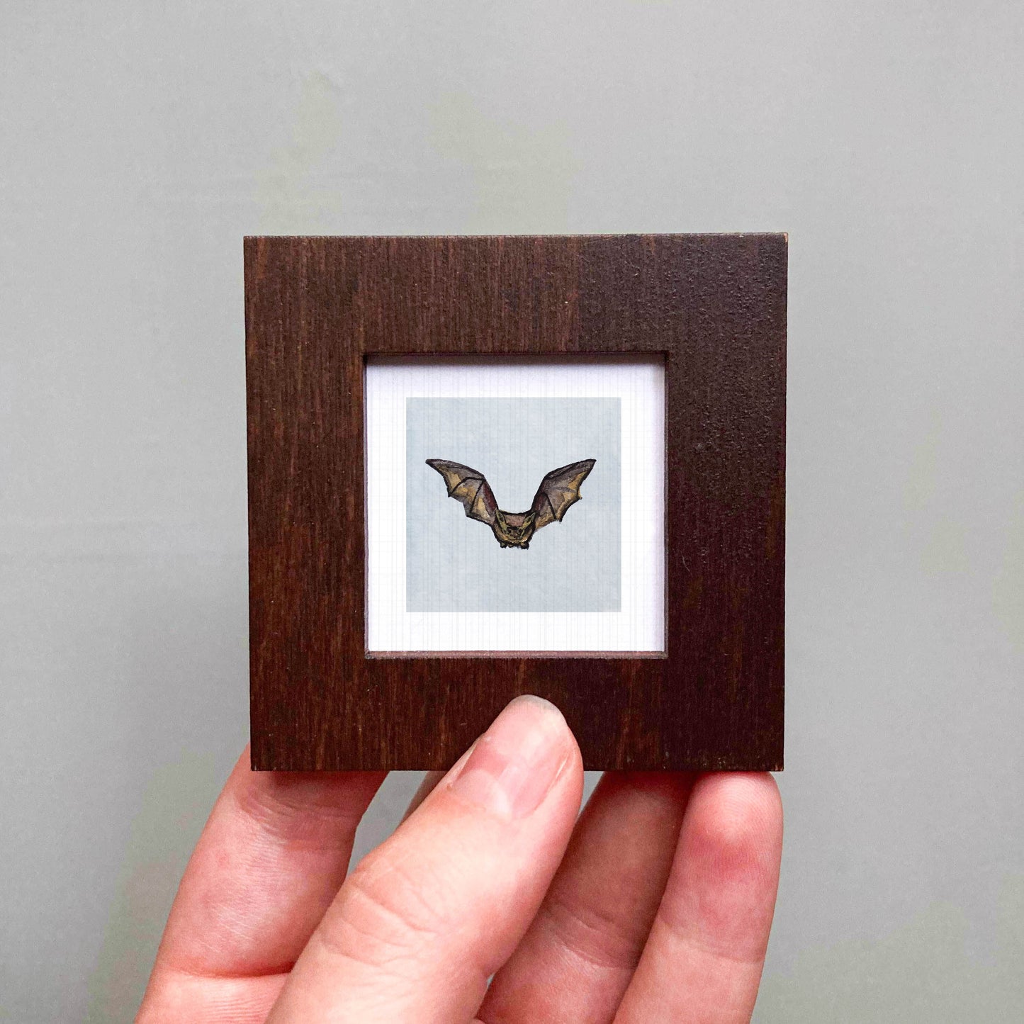 A person's hand is holding a small wooden frame containing a picture of a bat.